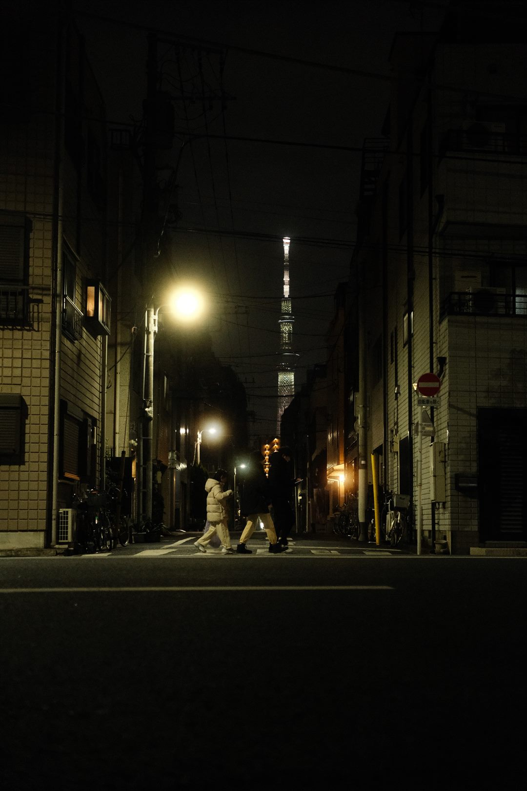 Tokyo Skytree lit at the end of a dark alley, three people crossing beneath a streetlight