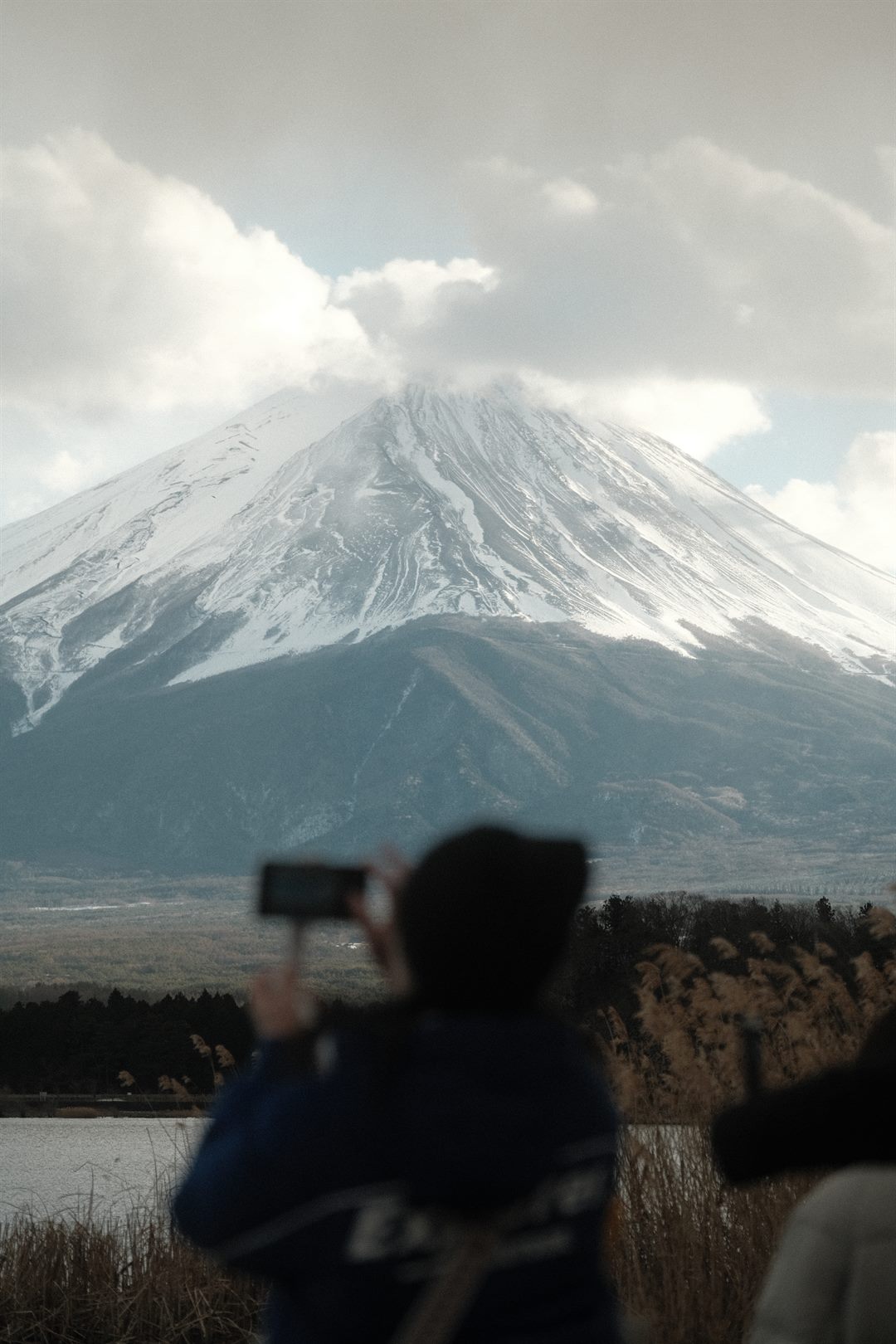 A person photographs Mount Fuji on a phone, their silhouette blurred in the foreground