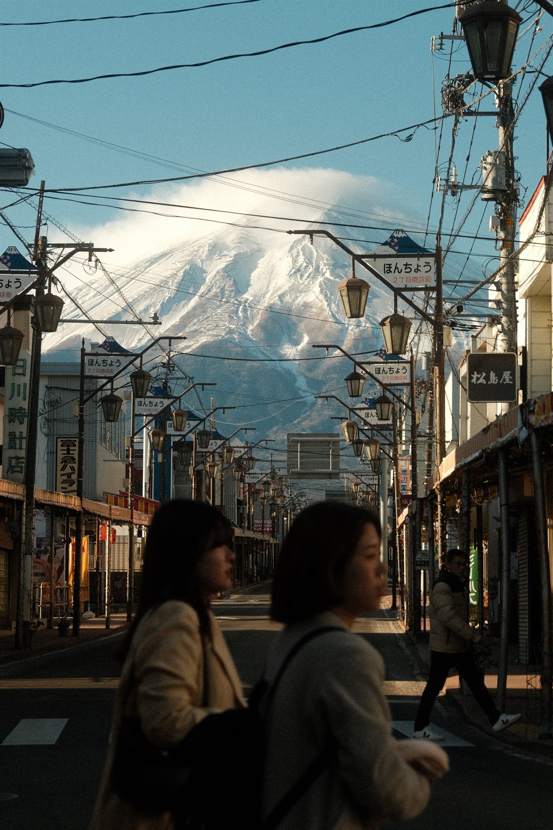 Two women cross a shopping street at dawn, Mount Fuji filling the far end