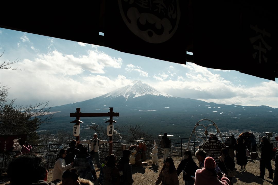 Crowds at a hilltop viewpoint under a dark shop awning, Mount Fuji across the valley