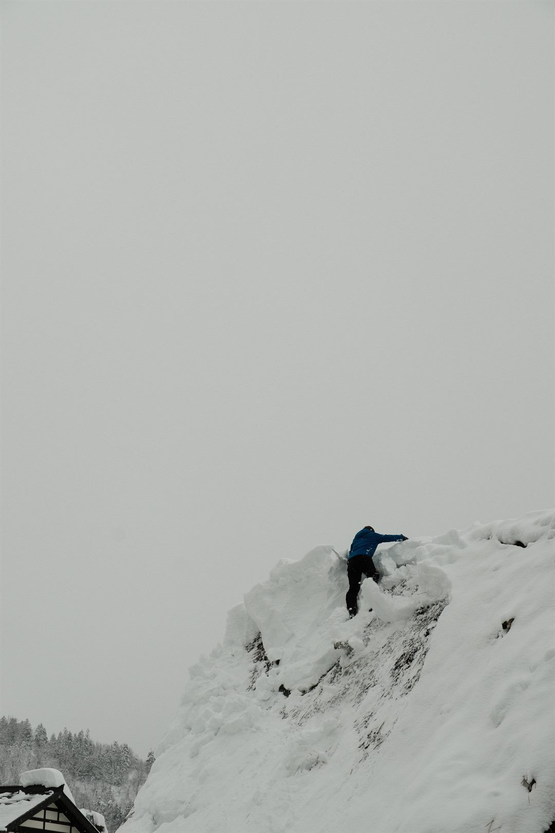 A person in a blue jacket clears snow from the top of a huge drift against a white sky