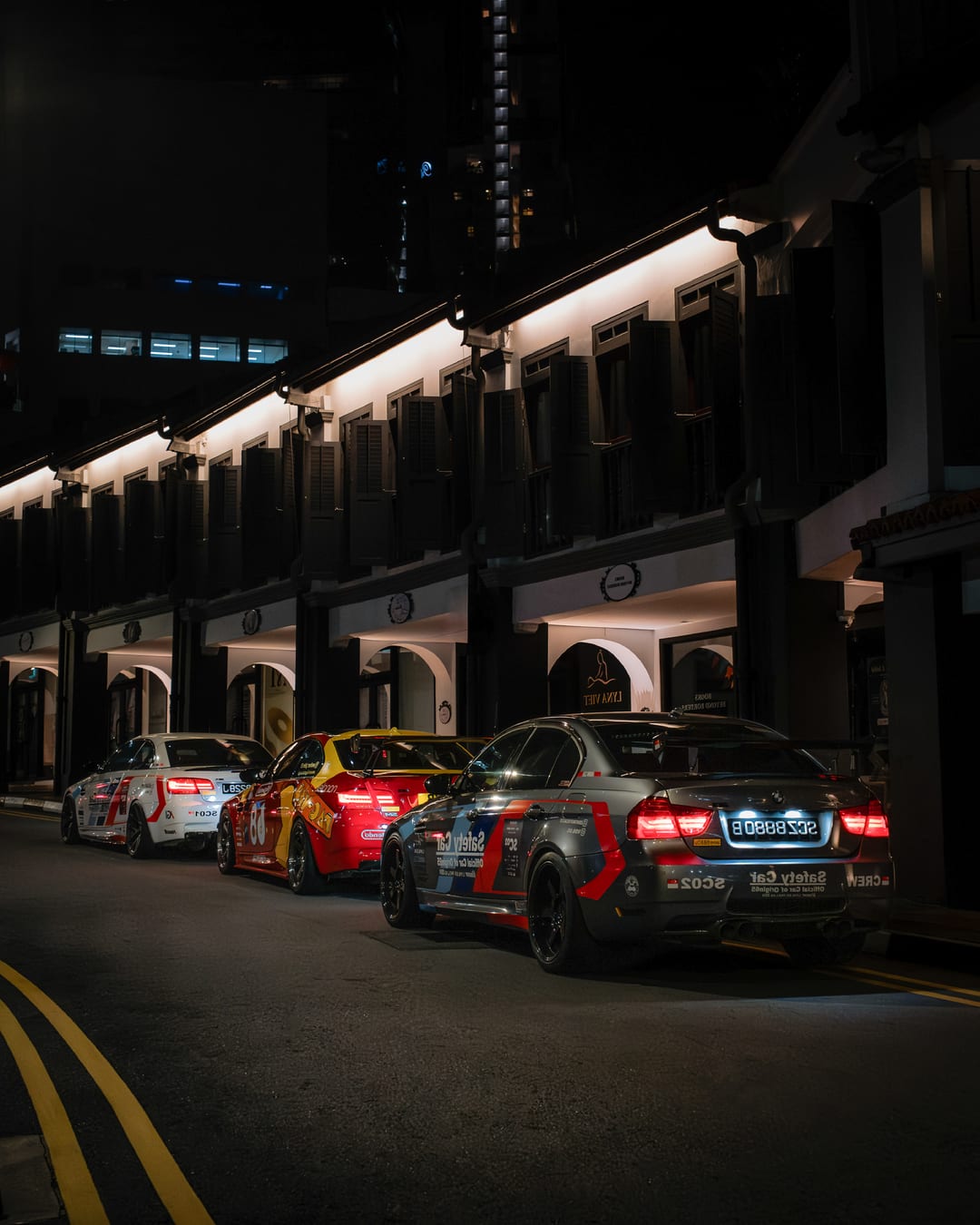 A Pac-Man liveried BMW M3 parked with other cars under a lit shophouse arcade at night